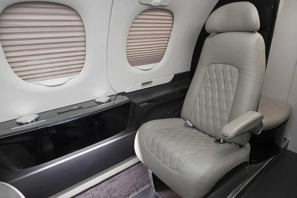 A light gray quilted leather passenger seat with a window and closed blinds in the cabin of a business jet.