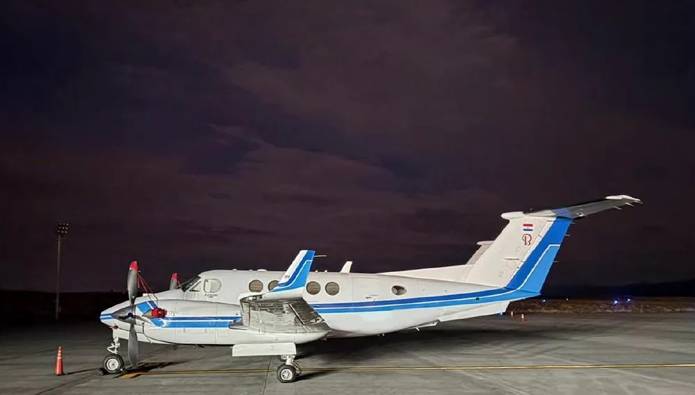 A white twin-turboprop aircraft with blue stripes and a flag on the tail, parked on an airfield at night.
