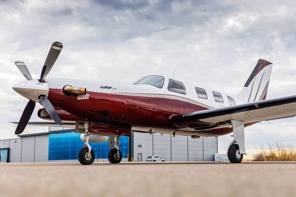A close-up view of the front-left of a white and maroon single-engine turboprop aircraft parked in front of a hangar.