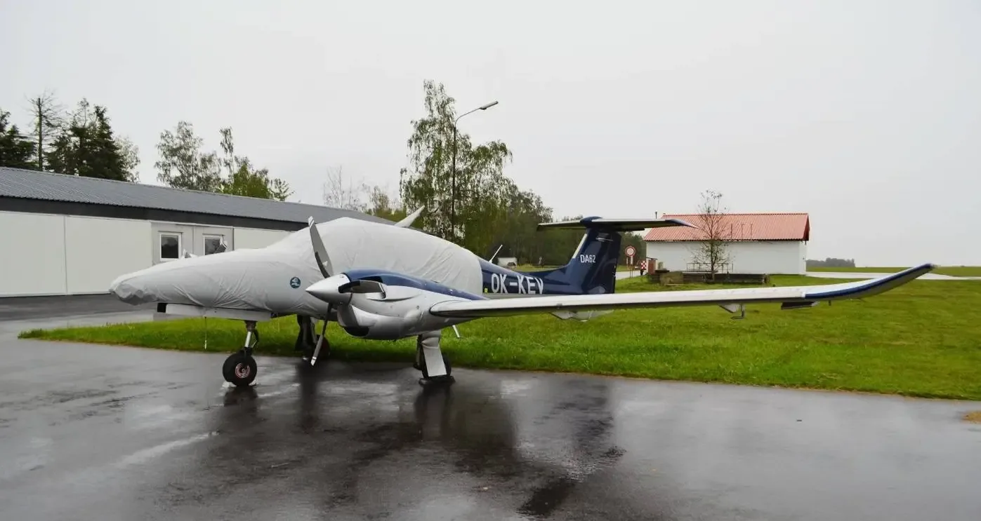 A blue and silver Diamond DA62 aircraft, partially covered, is parked on a wet tarmac next to a building.