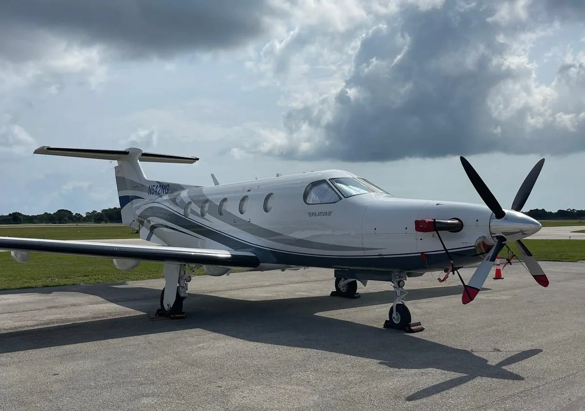 A white Pilatus PC-12 NG aircraft with blue and grey stripes, registration N542NG, parked on a tarmac with grass in the background.