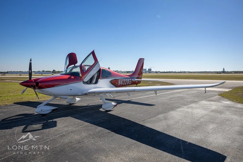 A front-side view of the red and white Cirrus SR20 aircraft (N432WB) with its doors open, parked on an airport tarmac.