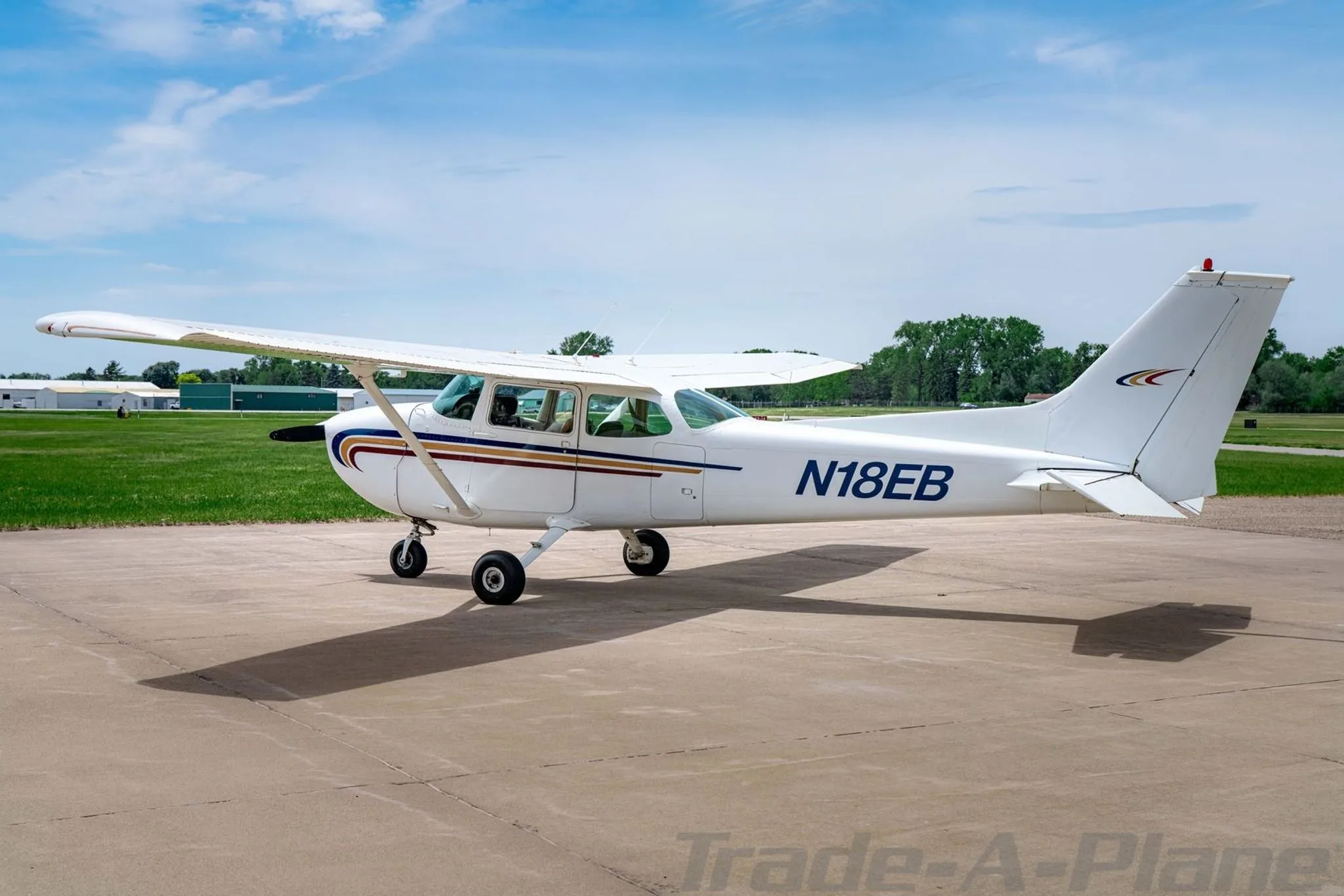 A white Cessna 172 with blue, gold, and red stripes on the fuselage, parked on an asphalt ramp, viewed from the rear-left.