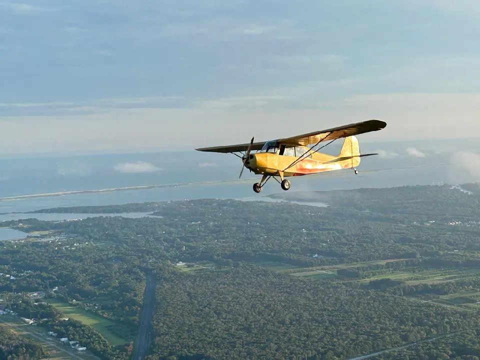 A yellow and orange Aeronca 7AC Champ aircraft flying over a green landscape with water in the background.
