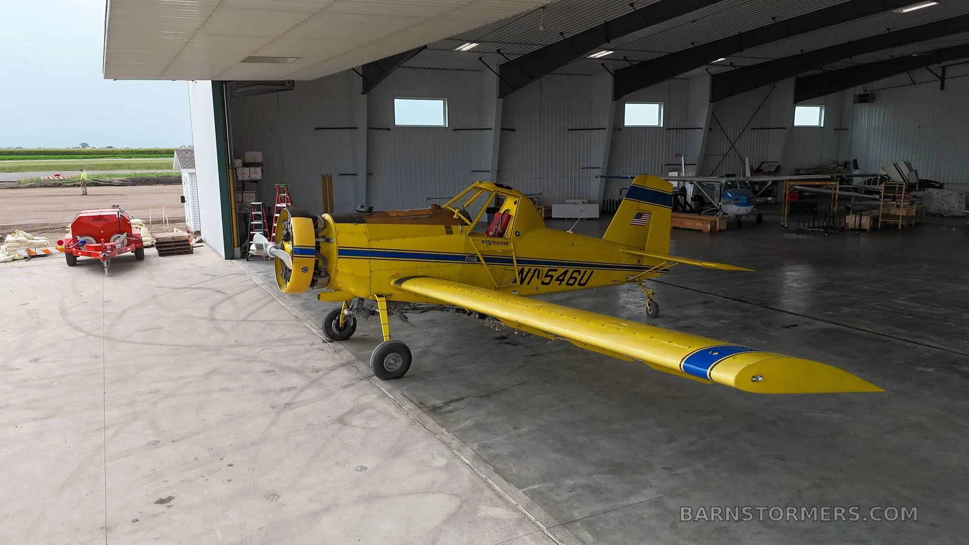 A yellow agricultural aircraft with blue stripes (N1546U) partially pulled out of a hangar, with a rural landscape visible outside.