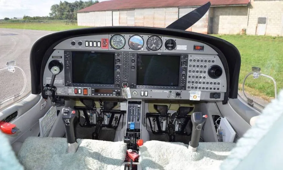 The cockpit of a Diamond DA40 aircraft, showing the instrument panel, controls, and light-colored seats.