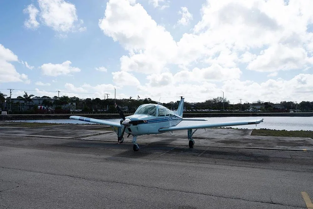 A light blue single-engine propeller aircraft, N9207S, is viewed from the front, parked on an asphalt apron with water and trees in the background.