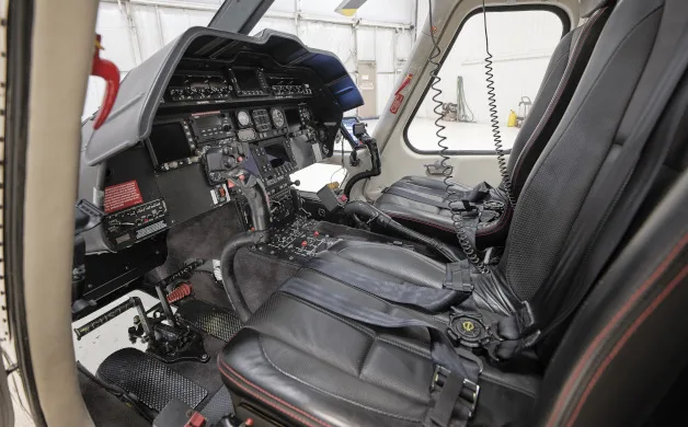 The cockpit of a helicopter, showing the instrument panel, controls, and black leather pilot seats from a side angle.