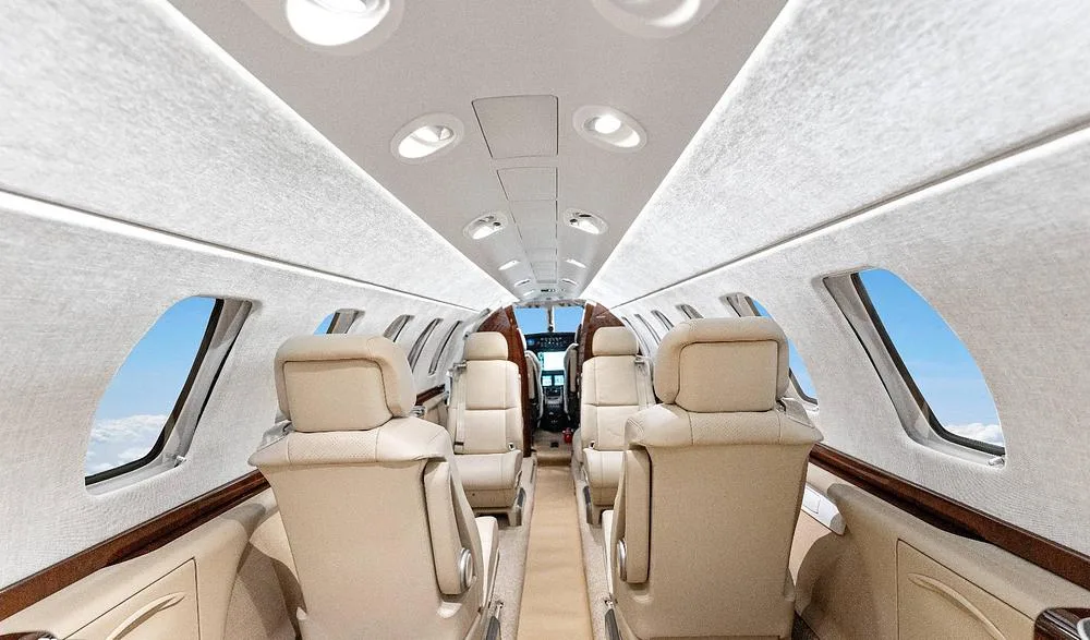 A long view down the aisle of a private jet cabin, showing multiple rows of beige leather seats and overhead lighting.