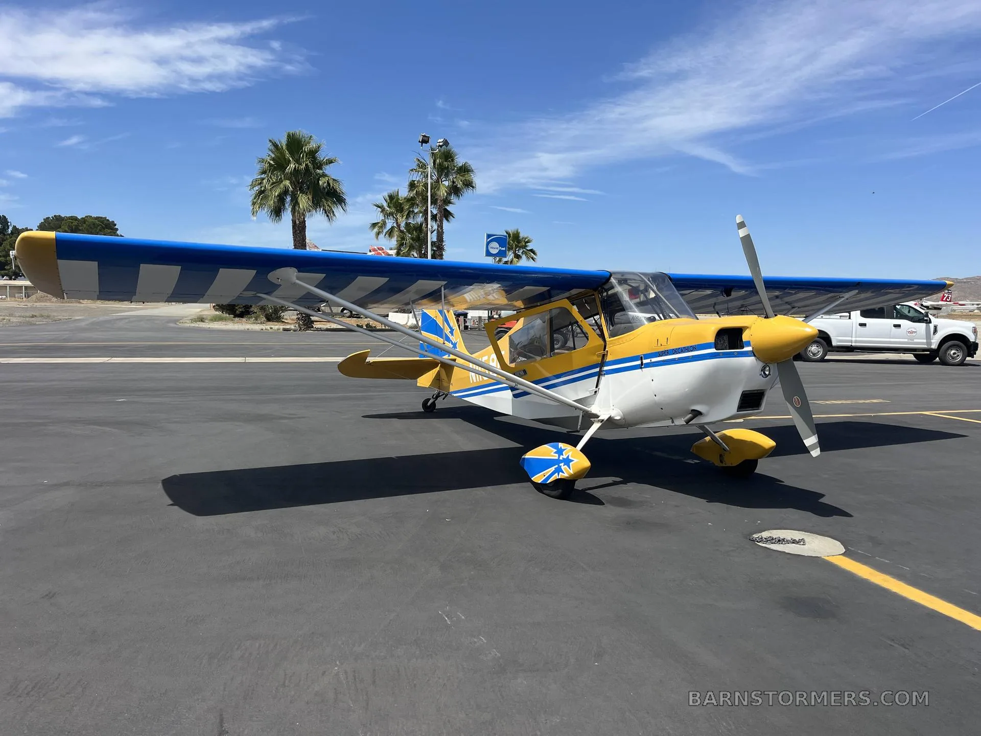 A yellow and blue high-wing aircraft with a striped wing and blue stars on the tail, parked on an asphalt tarmac under a blue sky with palm trees.