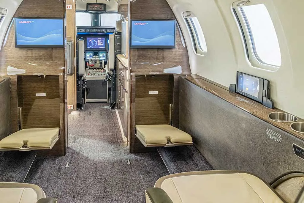 The forward section of the business jet's cabin, showing fold-down seats, entertainment screens, and a glimpse into the cockpit.