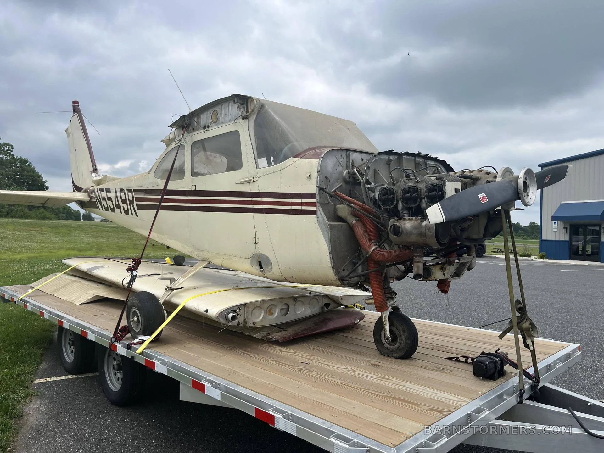 A partially disassembled white and maroon single-engine aircraft, registration N5549R, with its engine exposed, loaded on a trailer on an airfield tarmac.