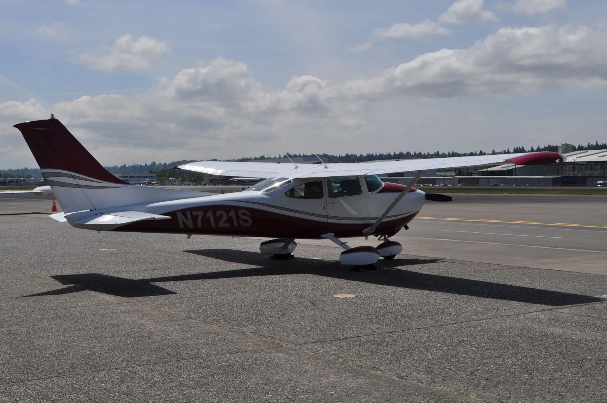 A red and white Cessna N7121S is parked on an airfield under a partly cloudy sky.