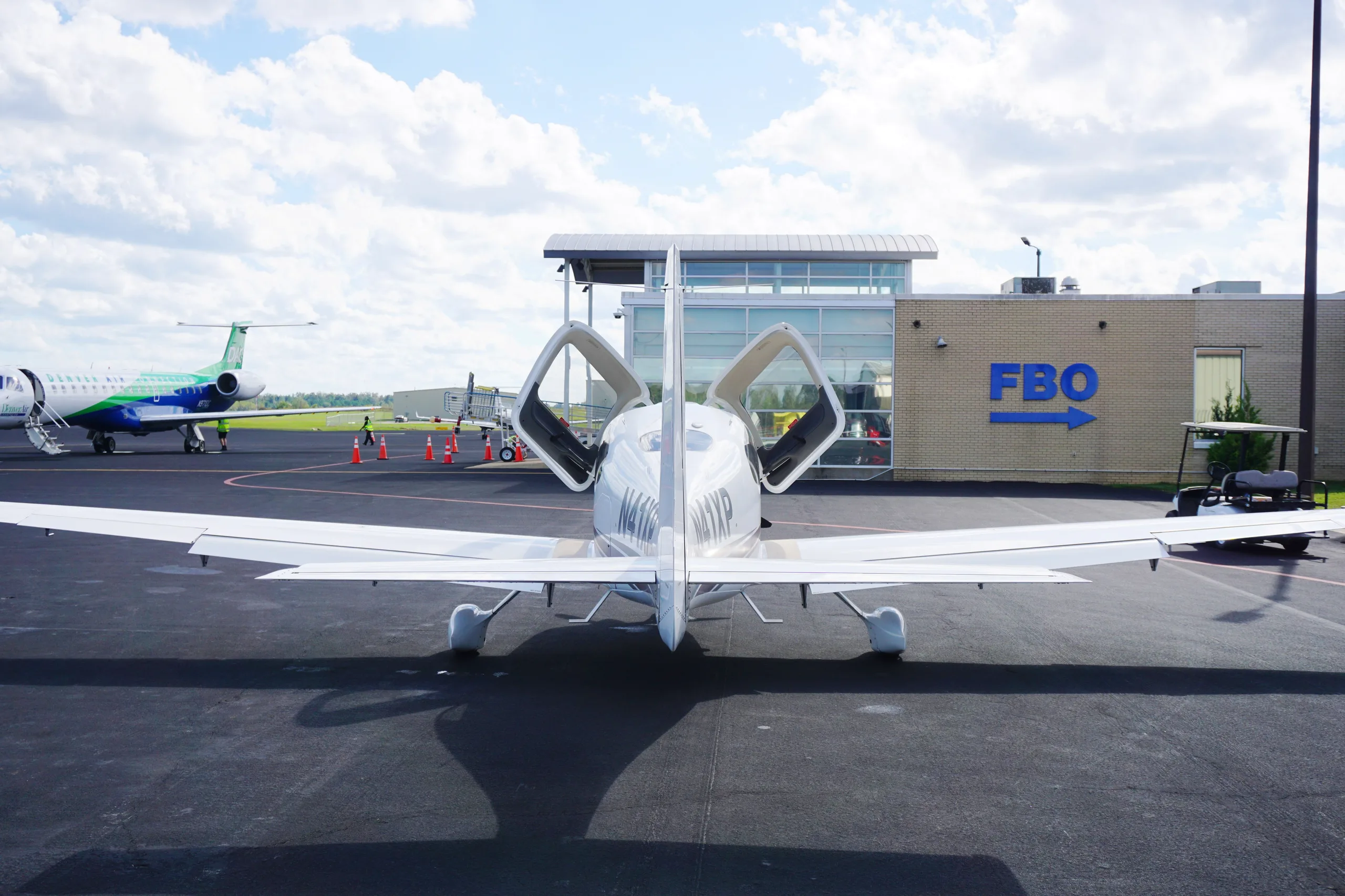 Rear view of a white Cirrus SR22 with its gull-wing doors open, parked on an airport tarmac in front of an FBO building.