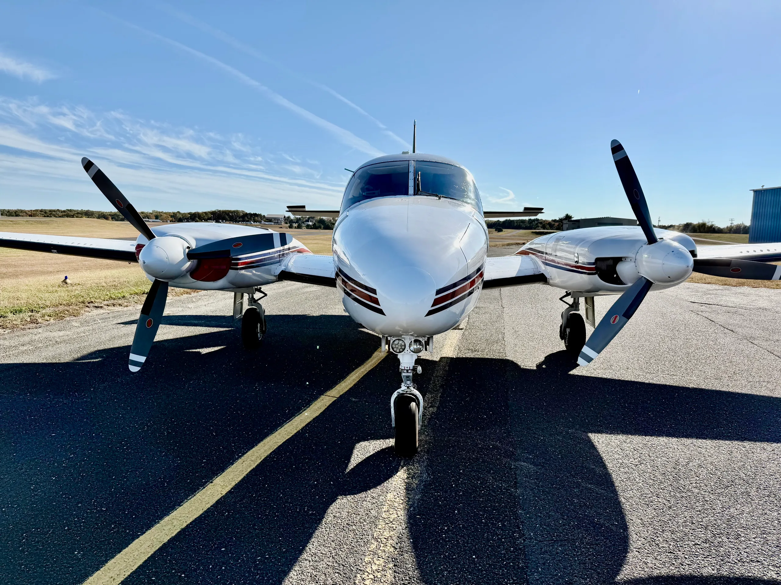 A head-on view of a white twin-engine propeller aircraft with red and blue stripes, parked on an asphalt ramp.