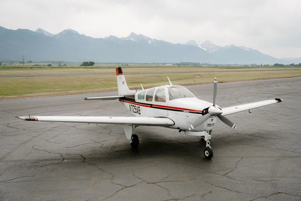 A white single-engine Beechcraft Bonanza aircraft with brown and orange stripes parked on a wet tarmac, with mountains and a runway in the background.