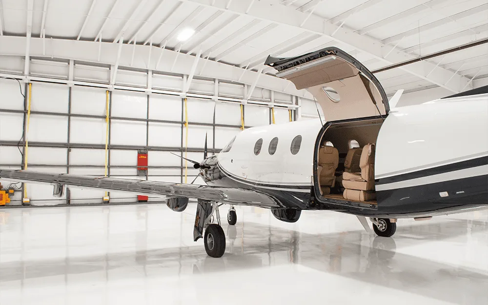 A black and white Pilatus PC-12 aircraft with its main cabin door open, revealing the interior, parked in a hangar.