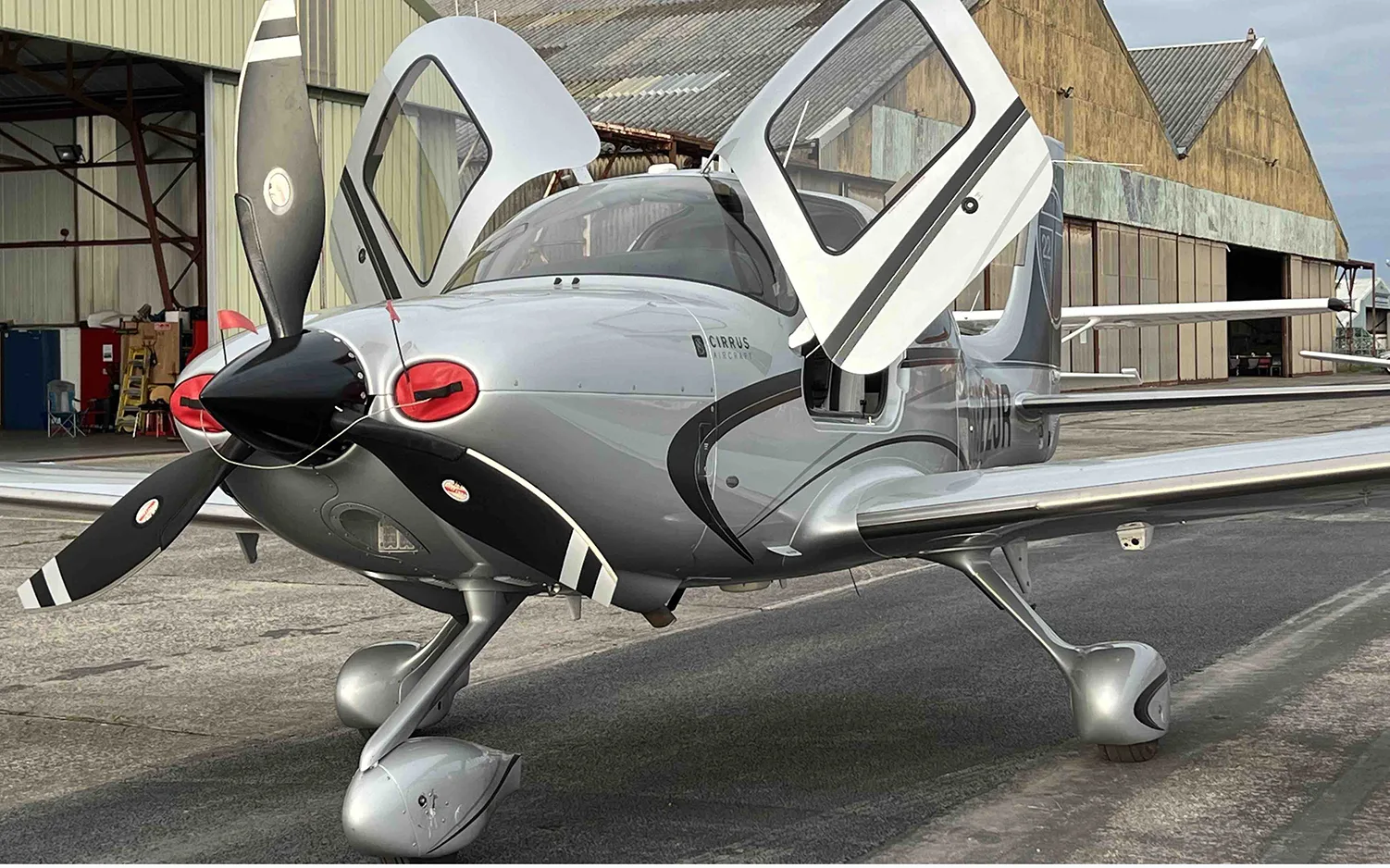 A silver Cirrus SR22 aircraft with black accents and open doors is parked in front of a hangar.