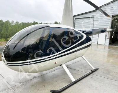 A white and blue Robinson R44 helicopter parked on a concrete pad next to a hangar on an overcast day.