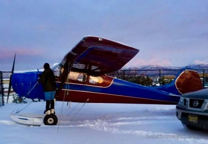 A red and blue high-wing aircraft on amphibious floats, parked in the snow next to a car, with a person standing on a float.