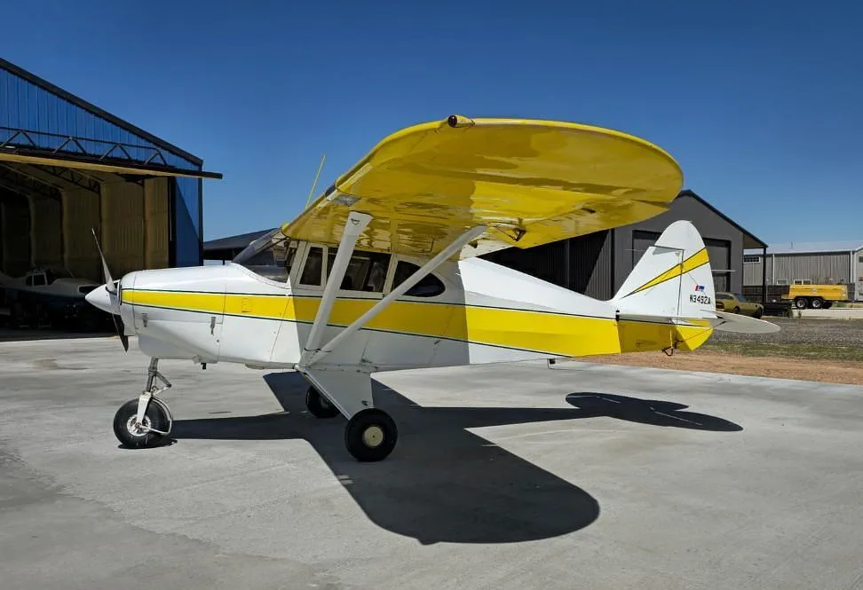 A side view of a white and yellow Piper PA-12 Super Cruiser (N3492A) parked on a concrete pad.