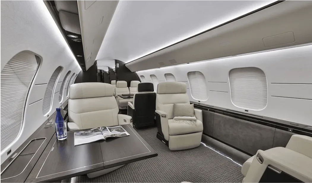 A wide view of a private jet cabin with light-colored leather seats, a table with a magazine, and large windows.
