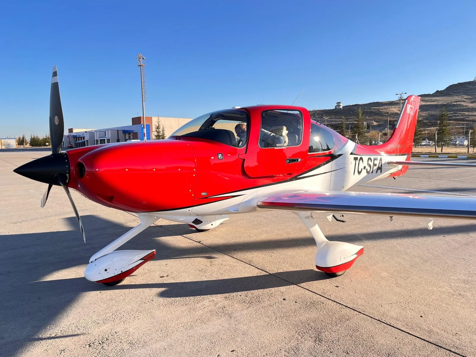 A red and white Cirrus SR22T aircraft with registration TC-SFA parked on an airfield under a clear blue sky.