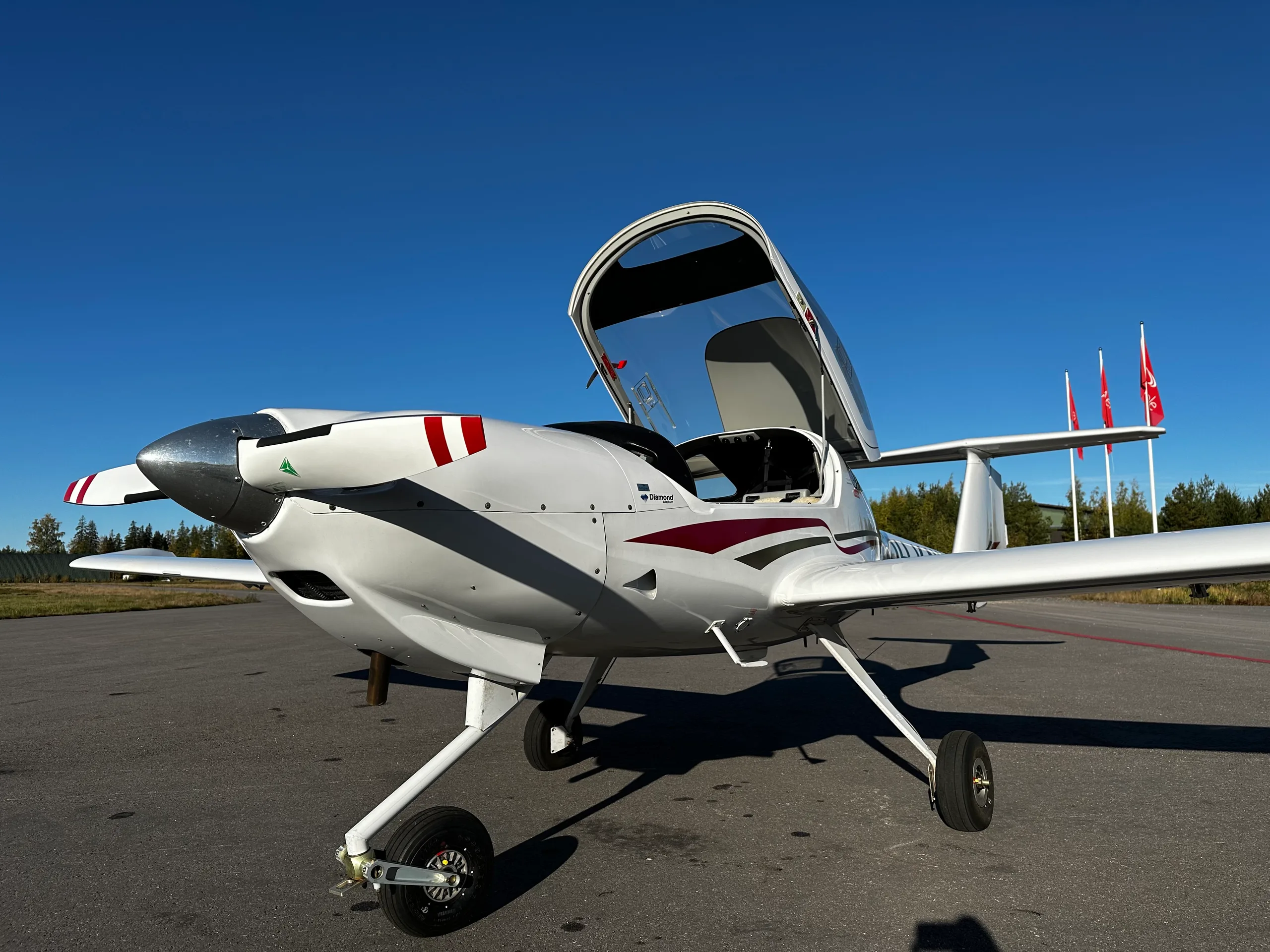 A white Diamond DA20 aircraft with its canopy open, showing red and black stripes on the nose, parked on an asphalt apron.