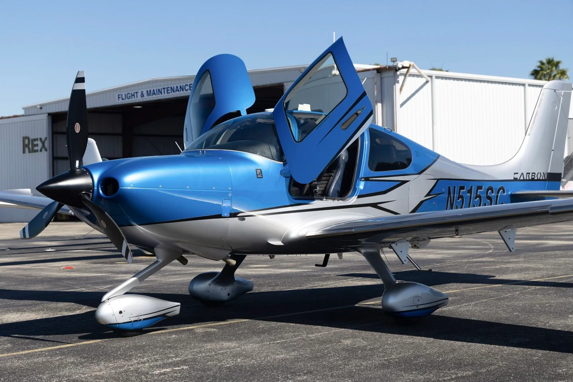 A blue and silver Cirrus SR22 aircraft (N515SC) with its doors open, parked on an airfield next to a hangar.