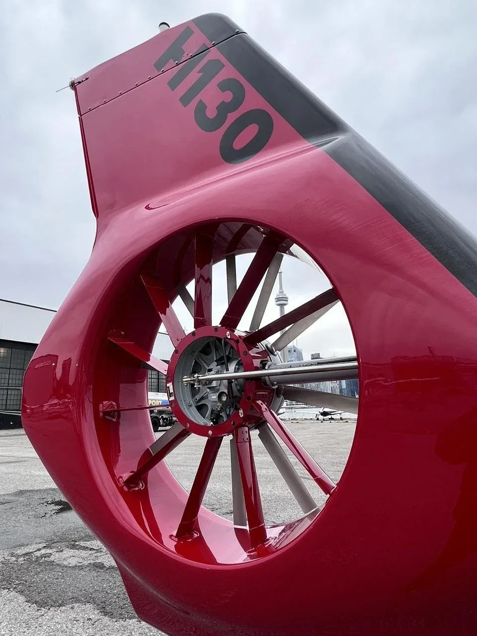 A close-up view of the red fenestron tail rotor of an Airbus H130 helicopter, with a city tower in the background.