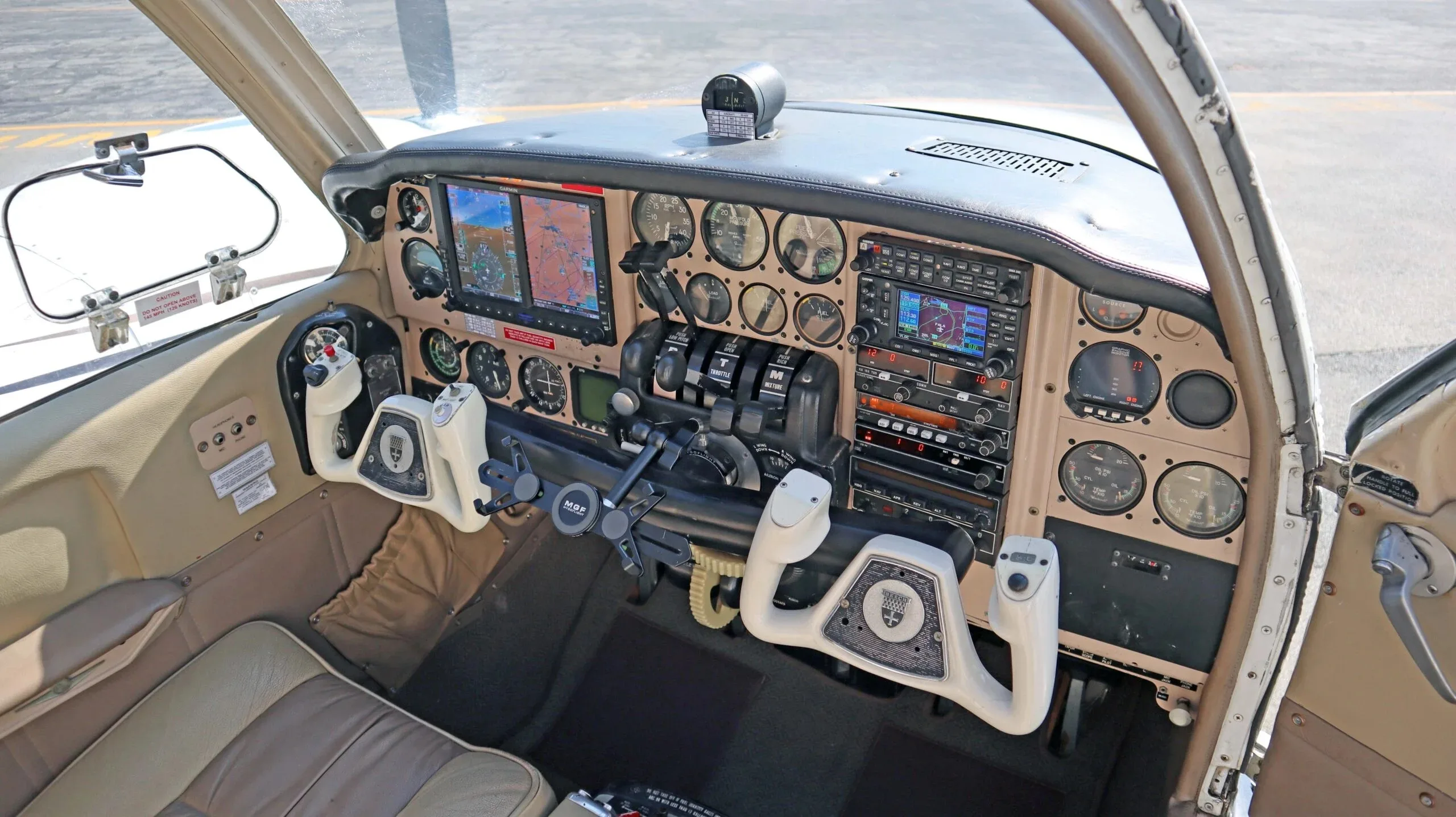 The cockpit of a small aircraft features a beige instrument panel with modern avionics screens and two white control yokes.