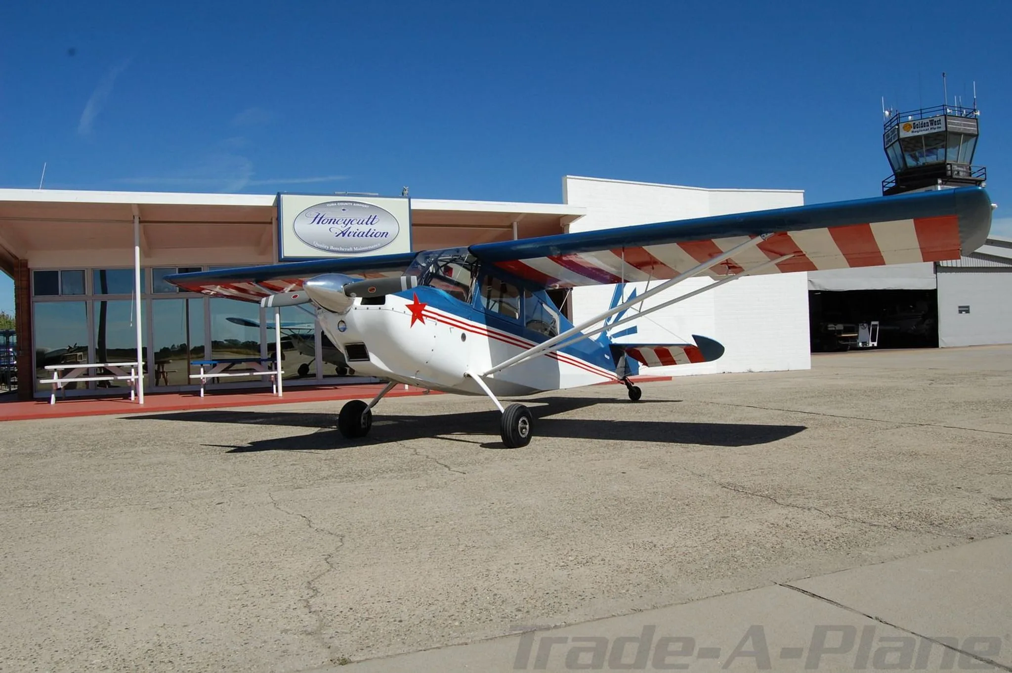 A blue, white, and red single-engine aircraft with a star on the nose, parked on an airfield next to a building with a 'Honeycutt Aviation' sign.