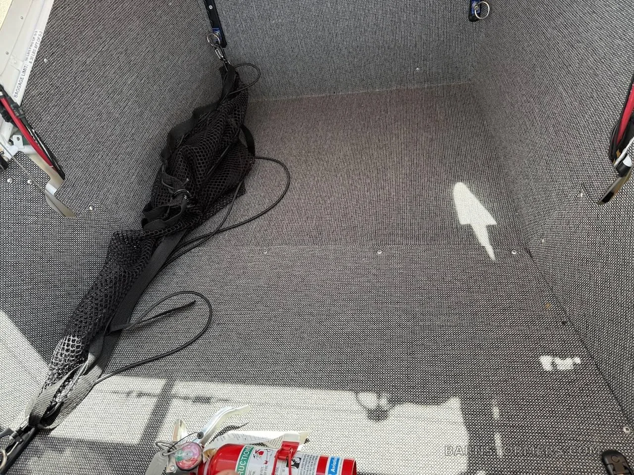The gray textured interior of an aircraft baggage compartment with a fire extinguisher and mesh bag.