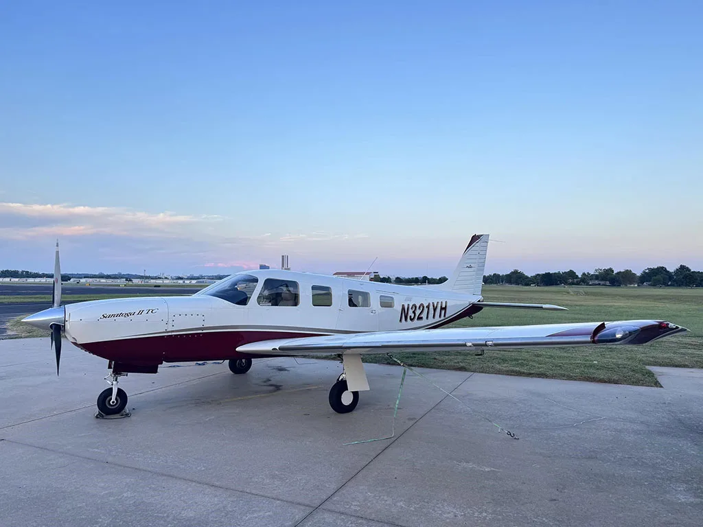 A white and maroon Piper Saratoga II TC aircraft (N321YH) tied down on an airport apron at sunrise or sunset.