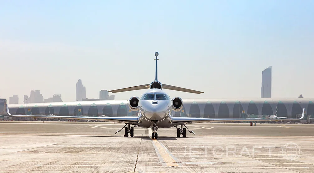 A white Dassault Falcon business jet with blue accents on the tail, seen from the front on a tarmac with a city skyline in the background.