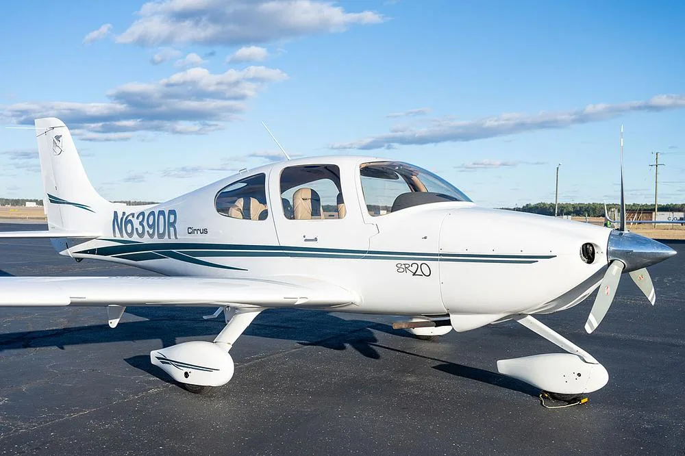 A white Cirrus SR20 aircraft with green and grey stripes, registration N639DR, parked on an asphalt tarmac under a blue sky.
