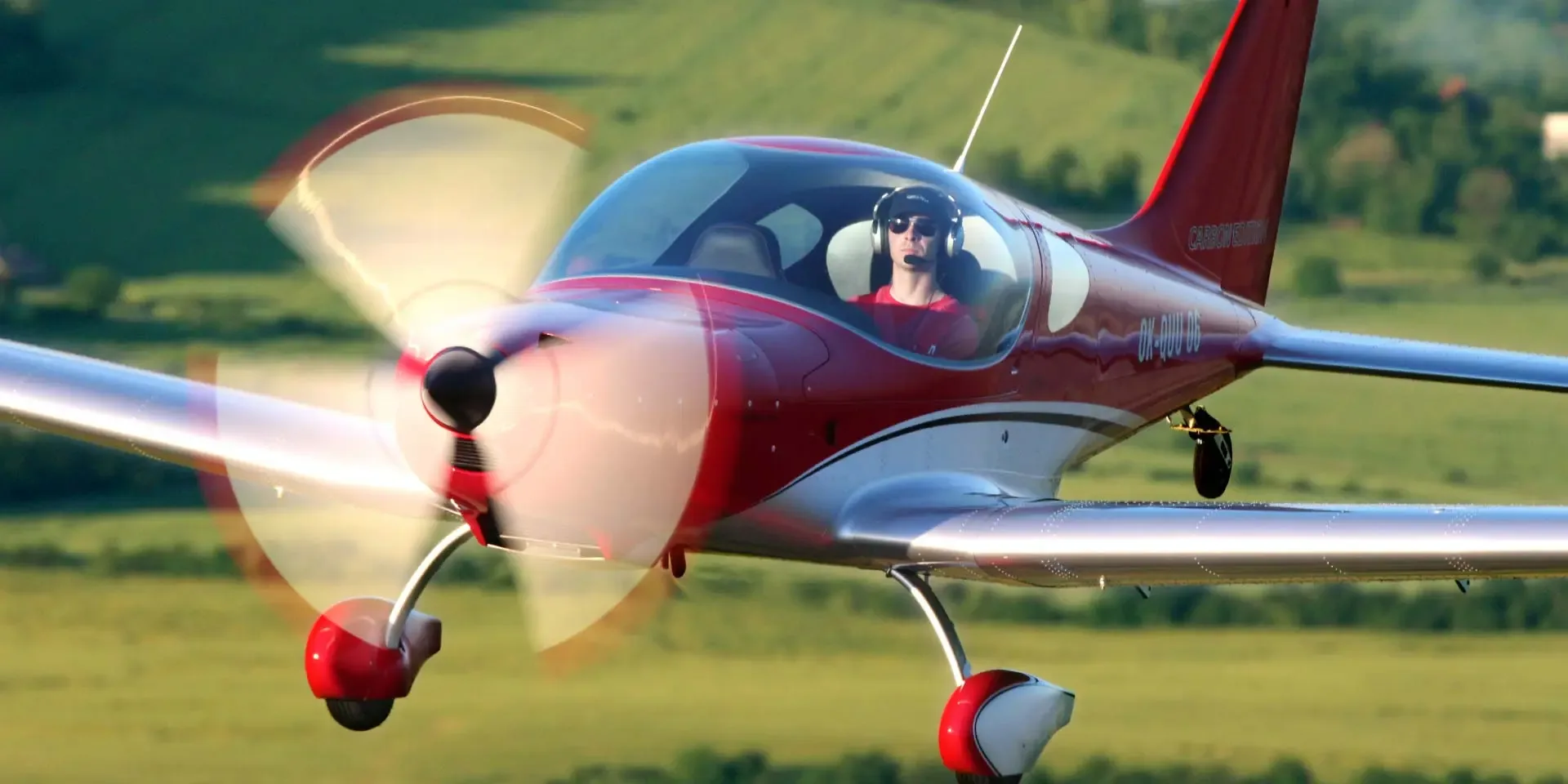 A red and white Carbon Cub aircraft with registration OK-QUU-06, piloted by a person wearing a headset, flying over a green landscape.