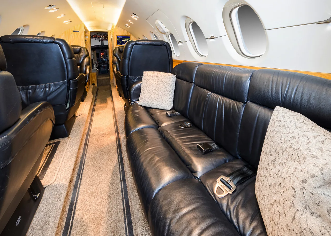 A view down the aisle of a private jet cabin with black leather seating and a glimpse of the cockpit.