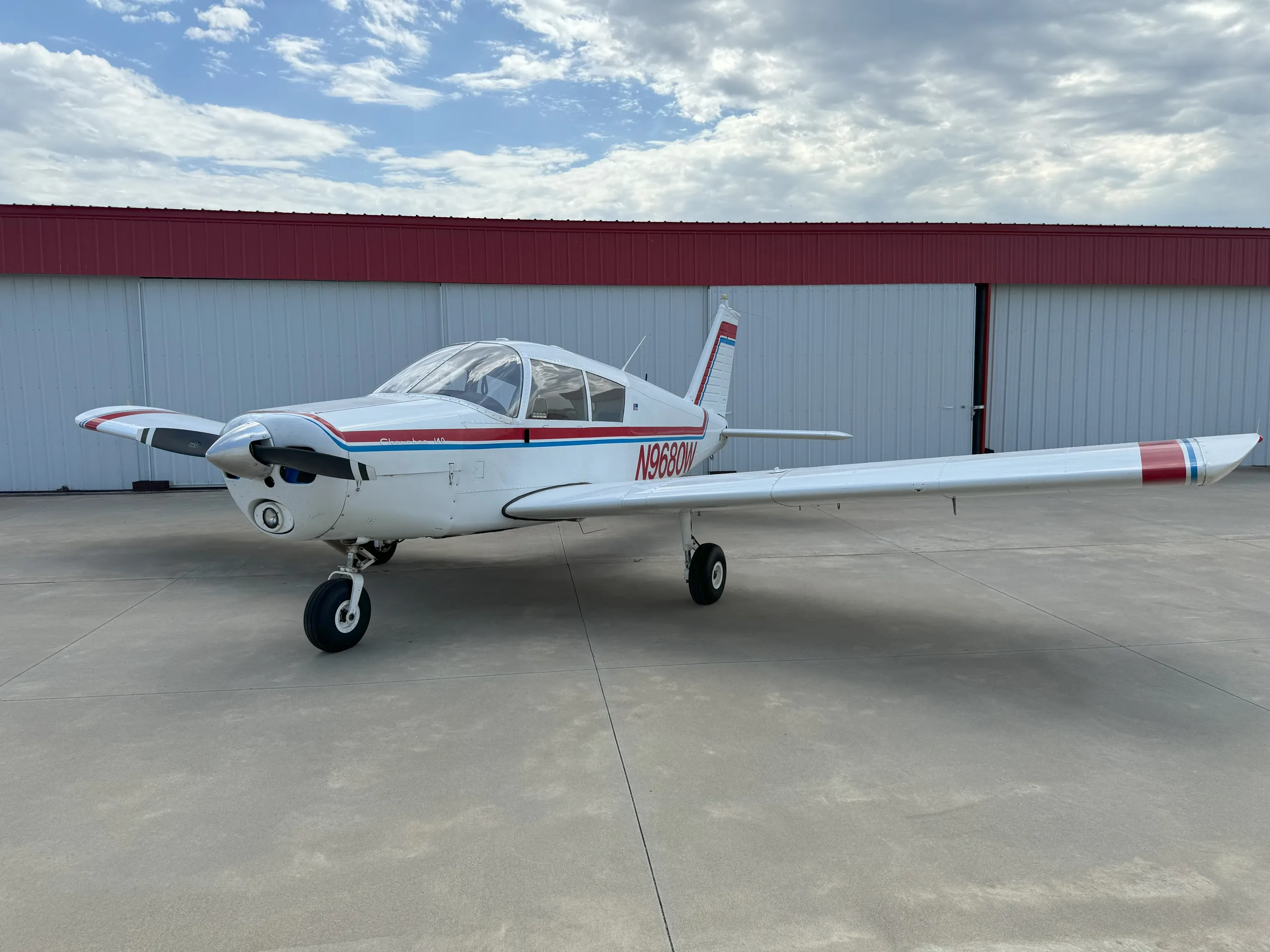 A white Piper Cherokee 140 aircraft with red and blue stripes is parked on a concrete tarmac in front of a hangar under a cloudy sky.