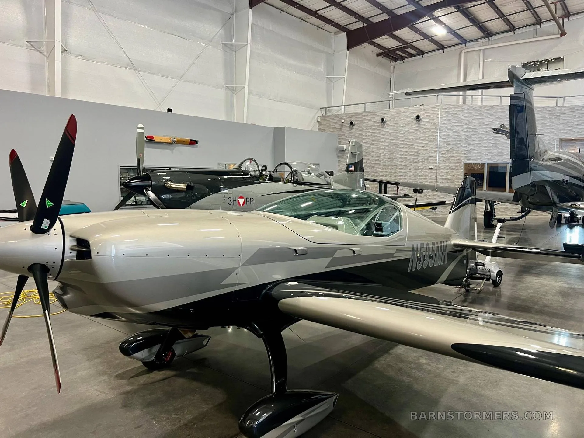 A front-side view of the silver and black propeller aircraft, registration N595MK, parked in a hangar with other aircraft.