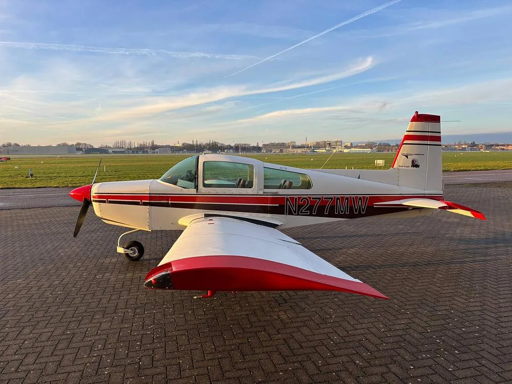 A white Grumman American aircraft with red and black stripes, registration N277MW, parked on a tarmac at sunset.