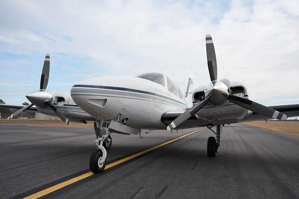 A low-angle front view of a white twin-engine propeller aircraft with blue and grey stripes on an asphalt surface.