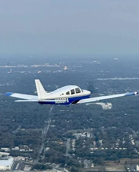 A white and blue Piper Arrow aircraft with registration N338PA flying over a city landscape with trees and buildings.