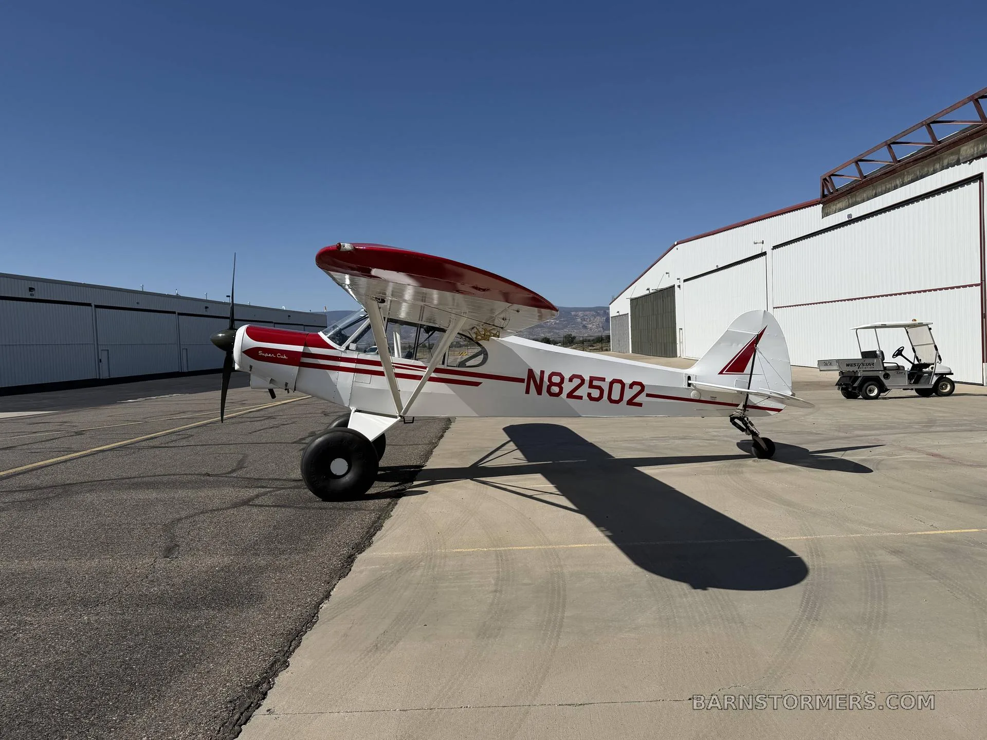 A white and red single-engine bush plane with large tires is parked on an asphalt apron next to a hangar, viewed from the side.