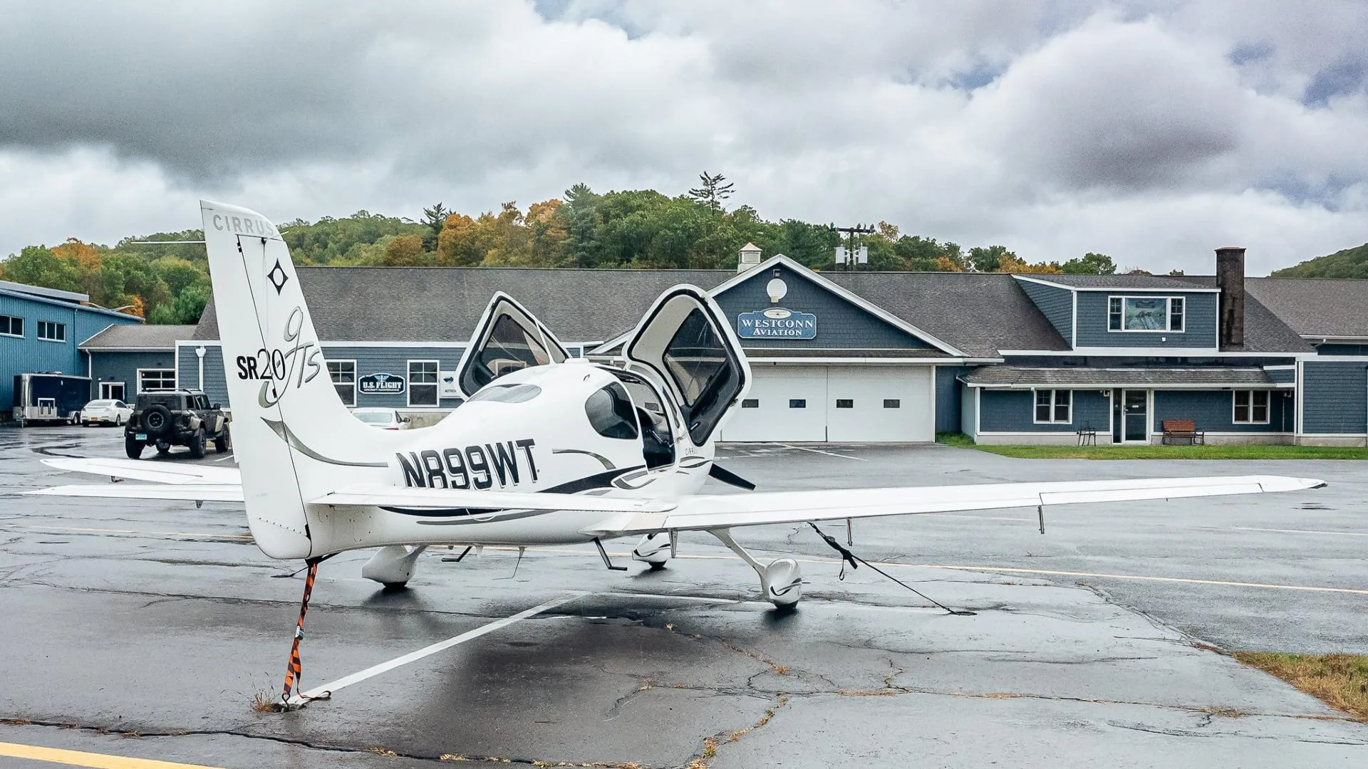 A white Cirrus SR20 aircraft with registration N899WT, parked on a wet tarmac with doors open, in front of a blue building.