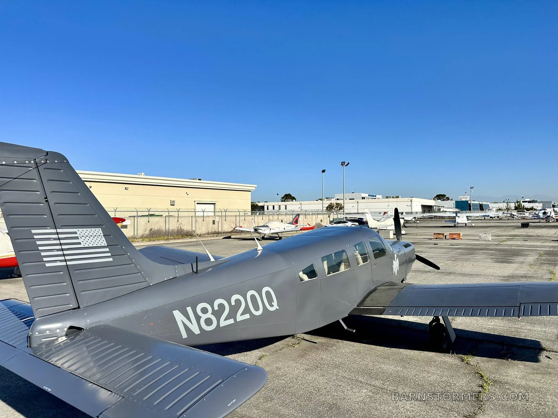 A rear-side view of a gray single-engine propeller aircraft, N8220Q, with an American flag on its tail, parked on a tarmac.