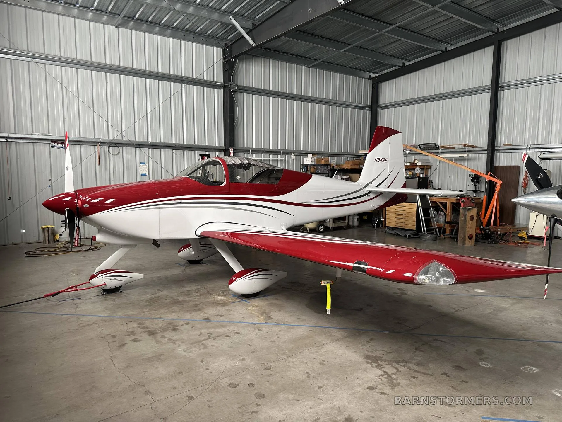 A red and white single-engine aircraft (N34BE) is parked inside a metal hangar.