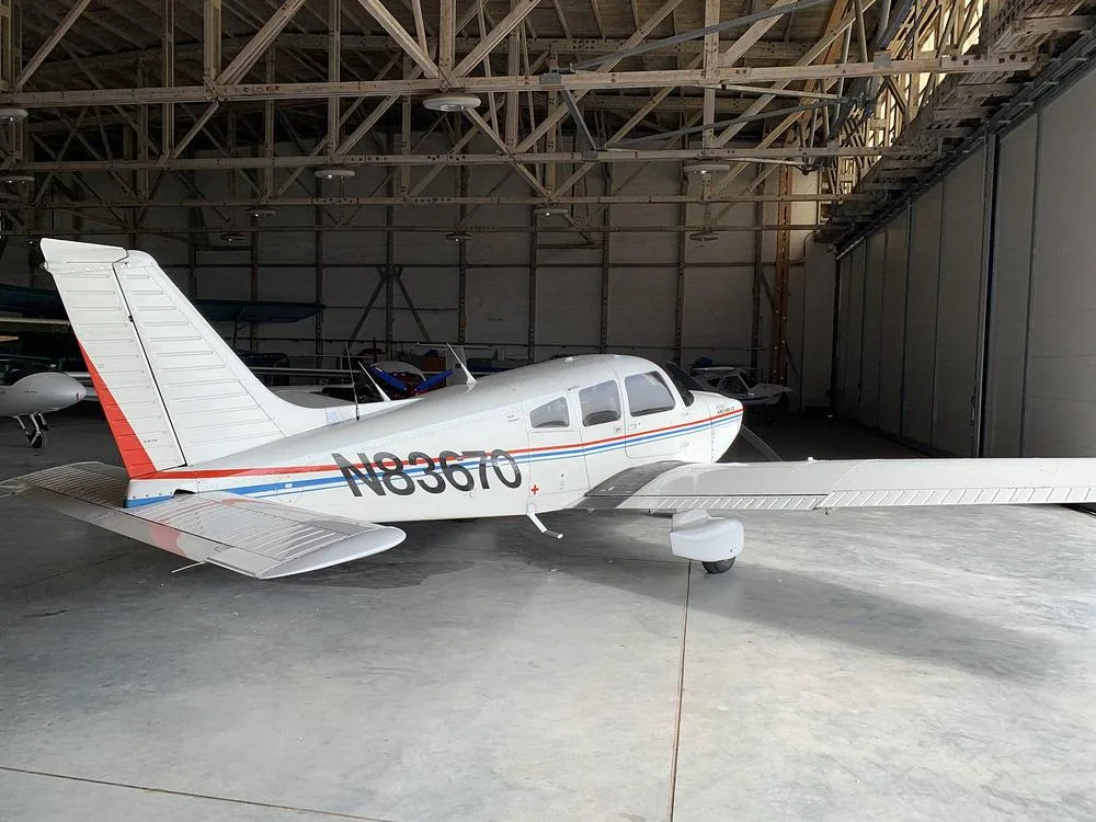 A white Piper Archer II aircraft with red and blue stripes parked inside a hangar.