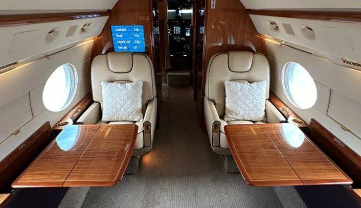 The luxurious interior cabin of a business jet with two beige leather seats facing each other, separated by wooden tables.