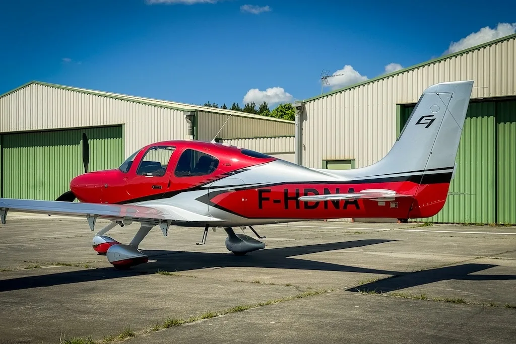 A red and silver Cirrus SR22 GTS (F-HDNA) aircraft parked on a tarmac in front of green hangars.